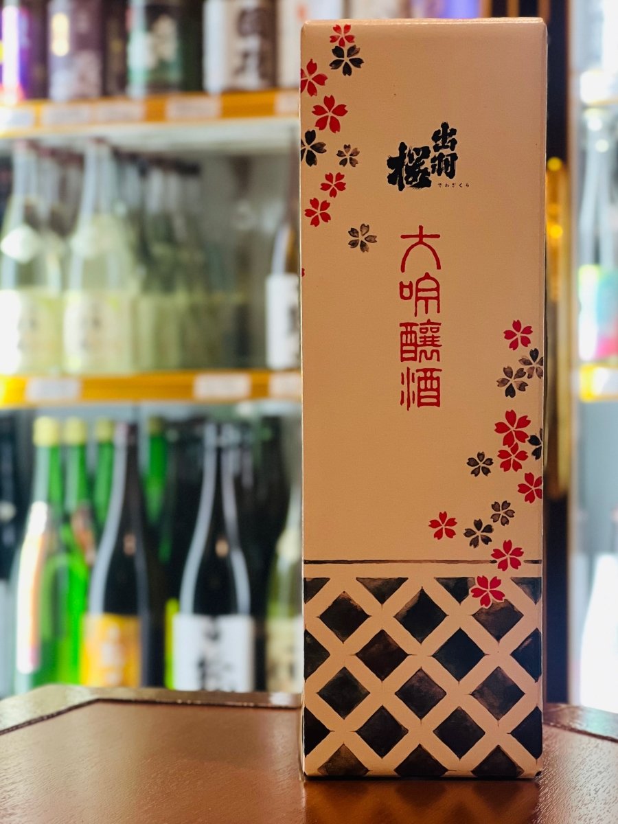 山形・出羽桜酒造の「出羽桜 大吟醸」720ml専用の化粧箱。温かみのある白を基調に、赤と黒の桜の花びらが舞う華やかなデザインが施されています。下部には伝統的な蔵を思わせる格子模様が配されており、日本初の市販大吟醸としての格式を感じさせます。バナナやカスタードのような優雅な香りを湛えた逸品を贈る、ギフトにふさわしいパッケージです。