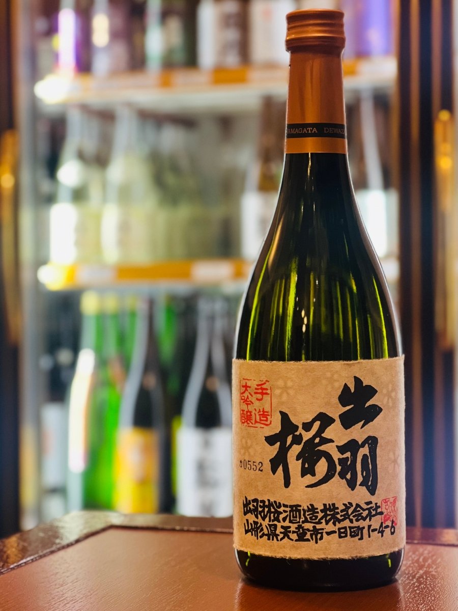 山形・出羽桜酒造の「出羽桜 大吟醸」720ml。冷蔵ショーケースを背に、木目調の台に置かれた濃緑色のボトル。耳付きの手すき和紙に墨書きされた伝統的なラベルが、繊細で優雅な味わいを物語ります。バナナやカスタードのような甘く華やかな香りと、氷温熟成によるまろやかな口当たりが楽しめる、吟醸酒の金字塔です。