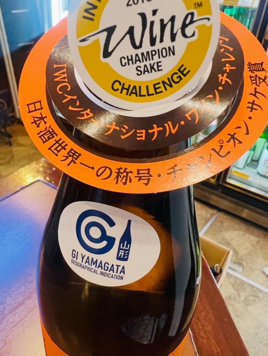 出羽桜「出羽の里」720mlのボトル首にかけられた、IWC（インターナショナル・ワイン・チャレンジ）の「チャンピオン・サケ」受賞を知らせる首かけラベル。オレンジ色の円形デザインに、大きく「Wine CHAMPION SAKE CHALLENGE」のロゴと、「日本酒世界一の称号」という力強いフレーズが記されています。ボトルの肩には「GI YAMAGATA」の認定ステッカーも貼られており、世界が認めた最高品質の純米酒であることを象徴する1枚です。