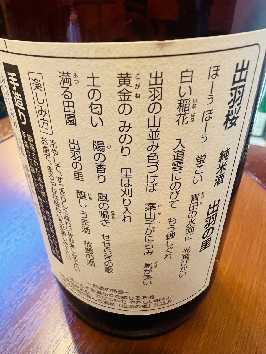 「出羽桜 純米酒 出羽の里」720mlの裏面ラベル。ラベルには、黄金の稲穂が実る山形の里山や、蛍が舞う美しい田園風景を詠った情緒的な詩が記されています。2016年IWCチャンピオン・サケ受賞の快挙を支える、厳選された米と蔵人の手造りへのこだわりを強調。「冷やしてすっきり、お燗でまろやか」といった、滑らかな口当たりを楽しむための案内が確認できる情報の接写画像です。