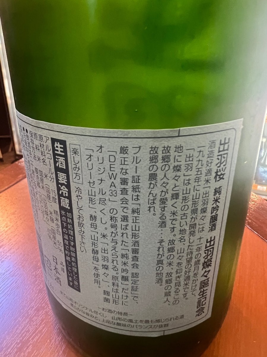 出羽桜「純米吟醸 出羽燦々誕生記念」の裏ラベル。山形県が11年かけて開発した酒米「出羽燦々」の由来や、山形オリジナル尽くし（米・麹・酵母）のこだわりが記されています。また、厳しい審査を通過した証である「DEWA33」の称号とブルー証紙（純正山形酒審査会認定証）についての説明、生酒の保存方法などが詳しく記載されています。