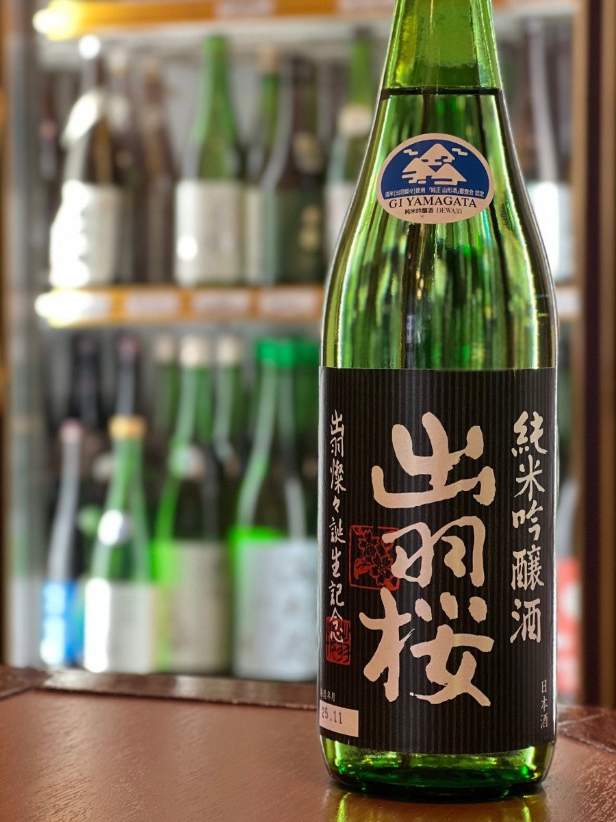 山形・出羽桜酒造の「純米吟醸 出羽燦々 誕生記念 火入」720ml。冷蔵ショーケースを背景に、黒いラベルに力強い金文字のロゴ、そして青い「GI YAMAGATA」の認定マークが輝くボトルです。山形県産の酒米、麹、酵母、水のみで醸された「オール山形」の一本。青リンゴのような爽やかな香りと、シルクのように滑らかな口当たり、上品な酸のバランスが絶妙な軽快食中酒です。