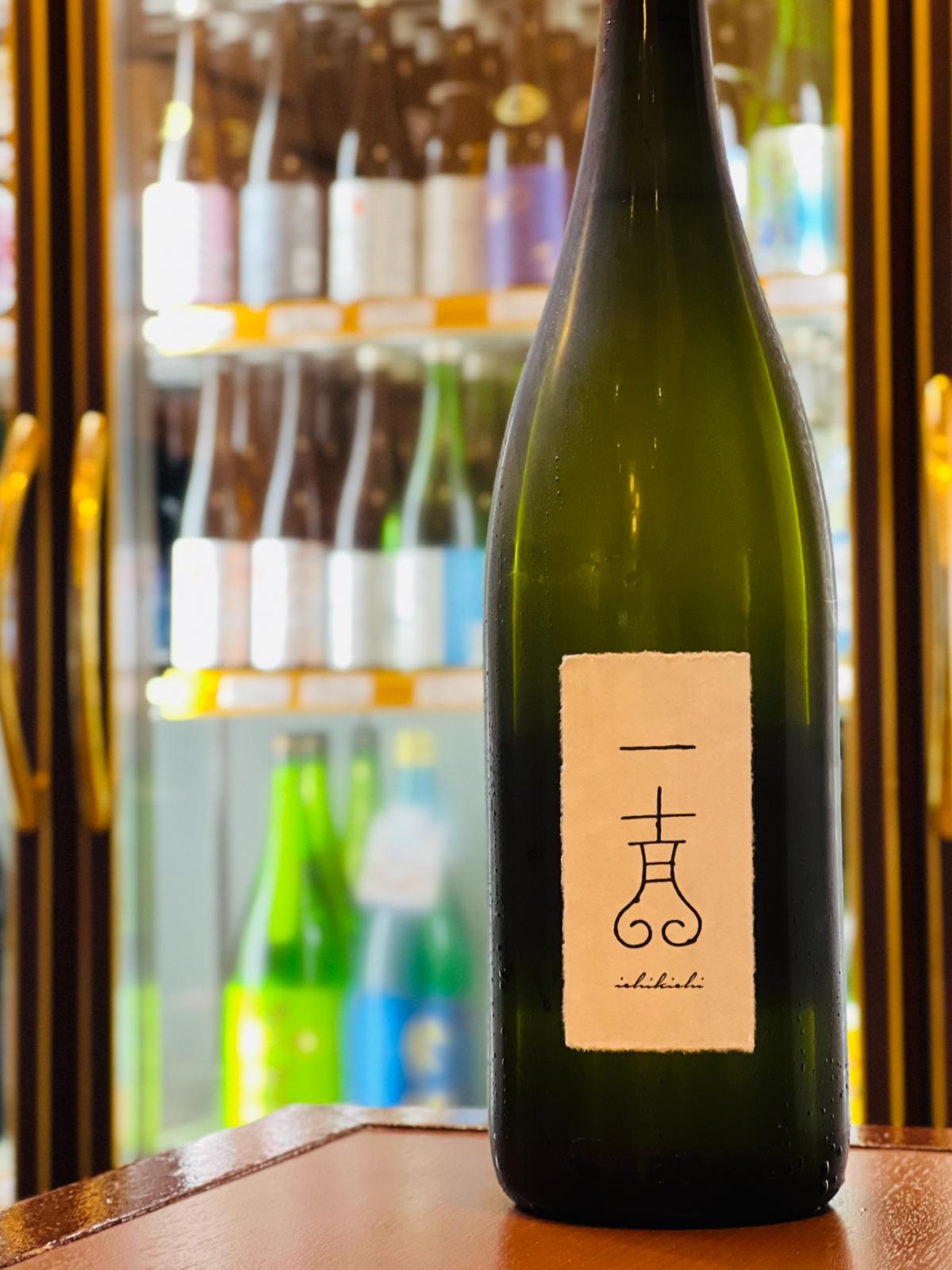 福島・国権酒造の「純米大吟醸 一吉（いちきち）1800ml」。冷蔵ショーケースを背景に、木目調のテーブルに凛と立つ濃緑色のボトル。清潔感のある白い和紙ラベルには、独創的な「一吉」のロゴと「ichikichi」の英字がシンプルに記されています。桃やメロンを思わせる瑞々しい香りと、超軟水仕込みによる「さらさら・ふわふわ」とした滑らかな口当たりが魅力の、上品で芯のある高級酒です。