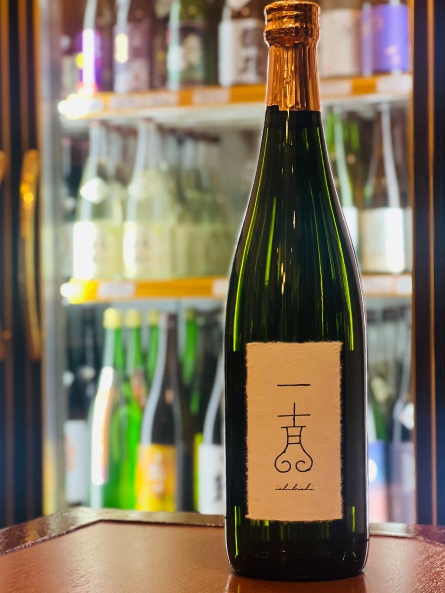 福島・国権酒造の「純米大吟醸 一吉（いちきち）」。冷蔵ショーケースを背景に、木目調のテーブルに凛と立つ濃緑色のボトル。清潔感のある白い和紙ラベルには、独創的な「一吉」のロゴと「ichikichi」の英字がシンプルに記されています。桃やメロンを思わせる瑞々しい香りと、超軟水仕込みによる「さらさら・ふわふわ」とした滑らかな口当たりが魅力の、上品で芯のある高級酒です。