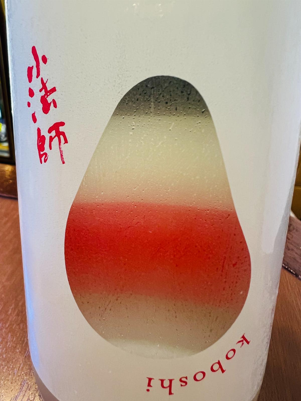 国権 純米酒 小法師のラベル接写。白地に赤字で「小法師」と記され、中央の小法師を思わせる形の一部が透明になっており、瓶の向こう側にある裏ラベルの色が透けて赤や淡い色のグラデーションとして見えるデザイン。