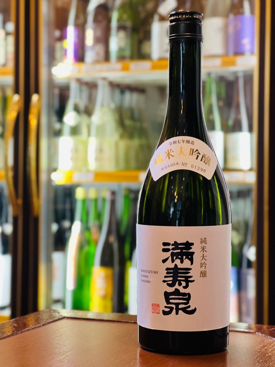 富山・桝田酒造店の「満寿泉 純米大吟醸」720ml。冷蔵ショーケースを背景に、木目調のテーブルに置かれた濃緑色のボトル。白い和紙ラベルに映える「満寿泉」の黒い筆文字と、醸造年度やシリアルナンバーが記された扇型の肩ラベルが目を引きます。シルクのような口当たりとバナナや梨を思わせるフレッシュな香りが魅力の、まさに「酒の芸術品」です。