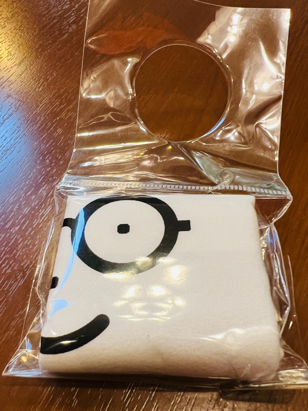 メガネ専用に付属するオリジナルのメガネ拭きが、ボトルの首に取り付けるパッケージへ折りたたまれている様子。メガネをかけた顔のデザインが印刷された、「メガネ専用」ならではの付属品。