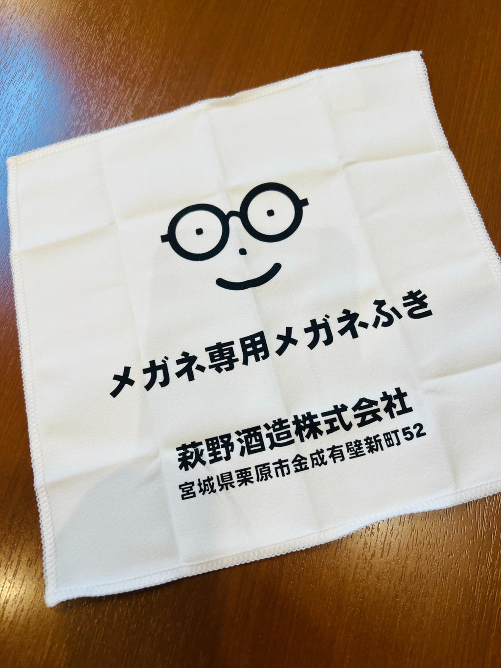 メガネ専用の付属品であるオリジナルメガネ拭きを広げた様子。白いクロスに、メガネの顔マークと「メガネ専用メガネふき」「萩野酒造株式会社 宮城県栗原市金成有壁新町52」の文字が印刷されている。
