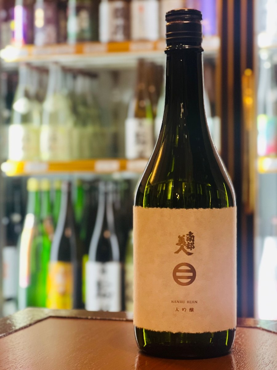 岩手・南部美人の定番高級酒「大吟醸」720ml。冷蔵ショーケースを背景に、木目調のテーブルに凛と立つ濃緑色のボトル。耳付き和紙のような質感のラベルには、伝統的なロゴと「NANBU BIJIN」の英字がシンプルに配されています。カスタードやバナナのような上品な甘い香りと、スターフルーツのような青い爽やかさが共存。しっとりと綺麗な酒質が魅力の、世界が認めた淡麗辛口な1本です。