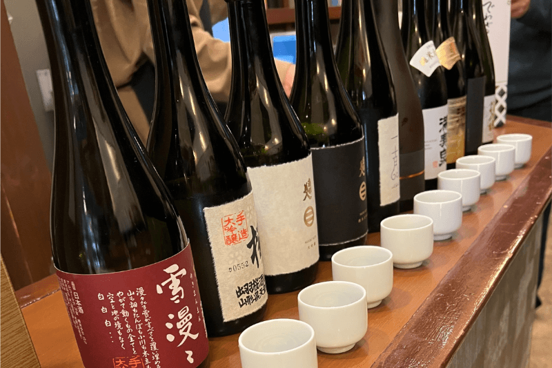 "出羽桜 雪漫々"、"満寿泉 寿"などがずらりと並ぶ。試飲会で高級日本酒を少量から試せる有料試飲。ギフト選びの際に味を確認できる店内の様子