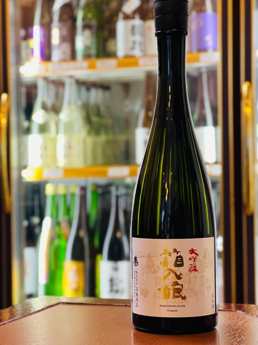 福岡・高橋商店の看板酒「繁桝 大吟醸 箱入娘」720ml。冷蔵ショーケースを背景に、スリムで気品ある黒いボトルが木目調の台に置かれています。ラベルには金色の箔押しで「箱入娘」の文字が美しく輝き、蜜の多いリンゴのような上品な香りと、さらさらと滑らかな口当たりを予感させる、洗練された佇まいの1本です。