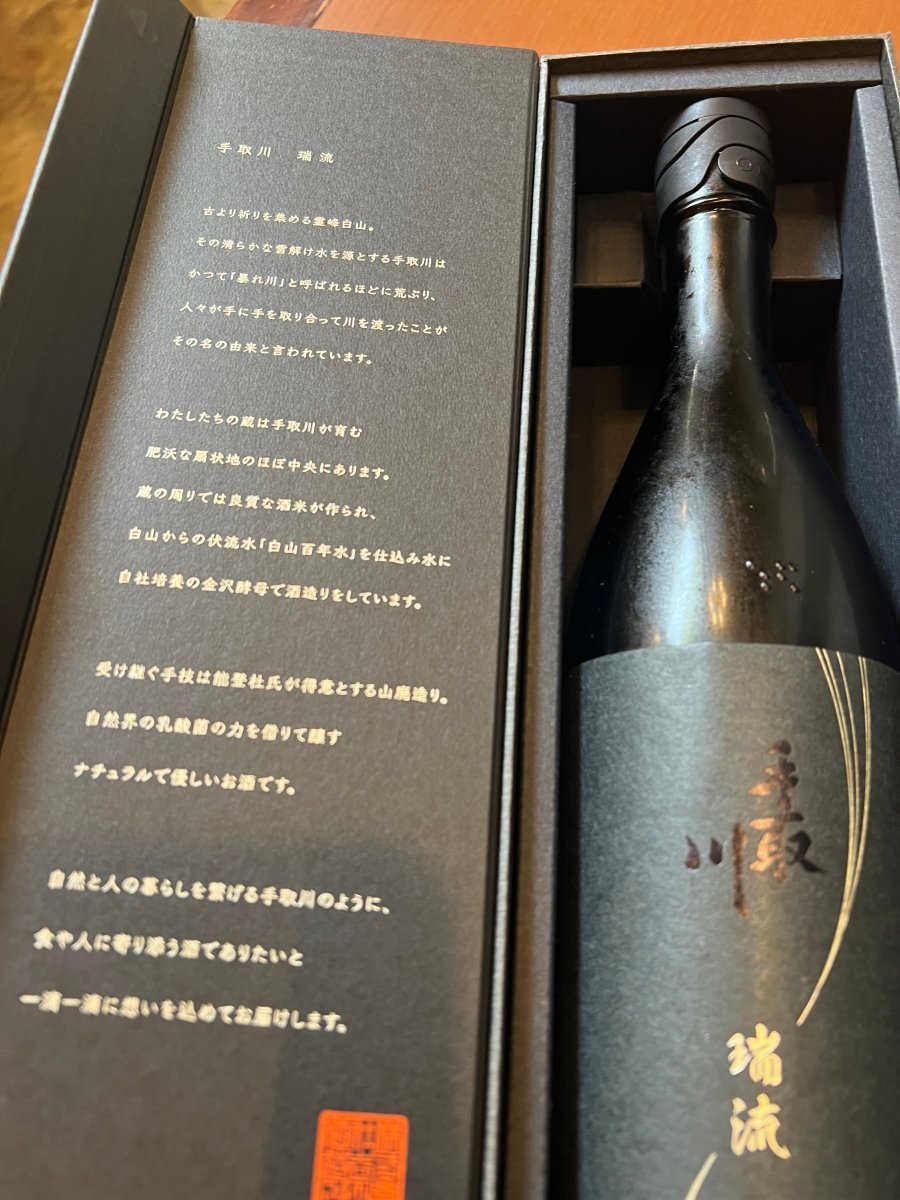 石川・吉田酒造店の最高峰「手取川 瑞流（ずいりゅう）」のギフト箱内部。内蓋には、霊峰白山の雪解け水を源とする「手取川」の名の由来や、肥沃な扇状地で育まれた良質な酒米、名水「白山百年水」への感謝が綴られています。自社培養の金沢酵母と、能登杜氏が得意とする伝統の山廃造りで醸された、一滴一滴に想いを込めたお酒の背景を伝える画像です。
