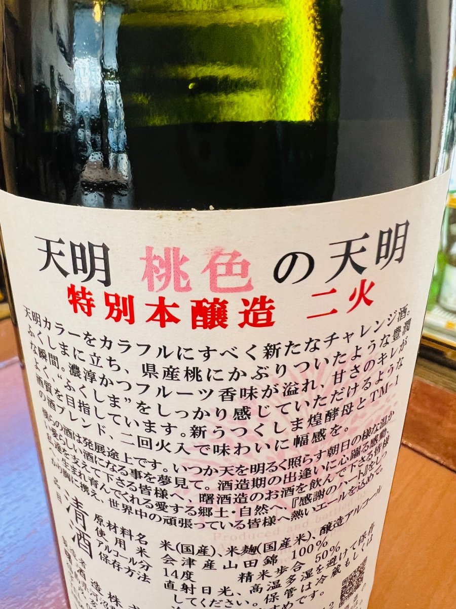 福島・曙酒造「桃色の天明（てんめい）特別本醸造 二火」の裏ラベル詳細。「天明カラーをカラフルにすべく新たなチャレンジ」として、県産桃にかぶりついたような豊潤な瞬間を表現した一歩です。新うつくしま煌酵母とTM-1のブレンド、二回火入れで味わいに幅を持たせています。「いつか天を明るく照らす朝日の様な温かい愛らしい酒に」という蔵元の夢と、愛する郷土・自然への感謝が綴られた、造り手の情熱が伝わるラベルです。