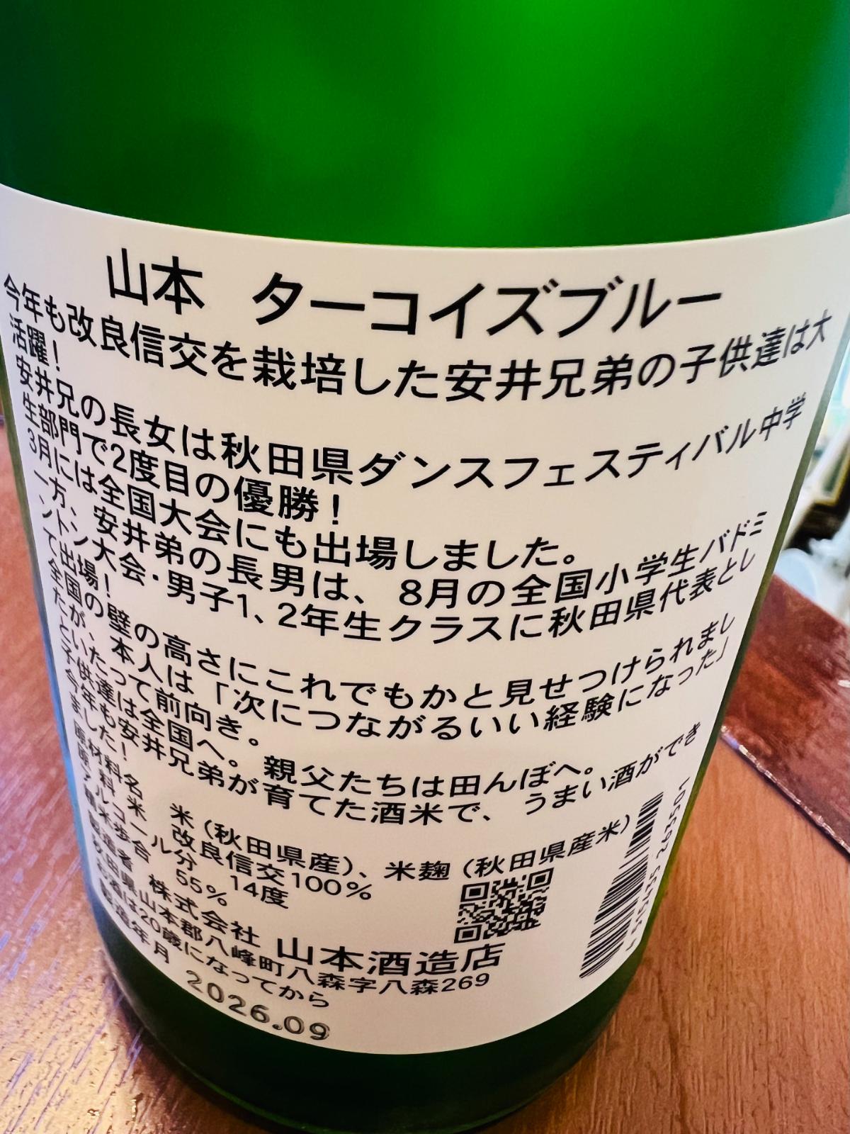 山本 純米吟醸 ターコイズブルーの裏面ラベル。酒米「改良信交」を栽培する安井兄弟の子どもたちの近況が毎年紹介される特徴的なラベルで、2026年はダンス大会やバドミントン大会での活躍など、栽培農家の家族の成長が記されている。原料米は秋田県産改良信交100％、精米歩合55％、アルコール分14度。
